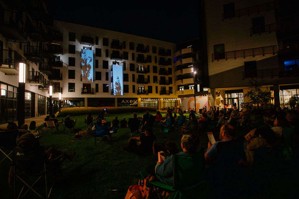 Opera Columbus performers singing from balconies at Gravity while OBLSK projection-mapped visuals onto flowing material, with hundreds of attendees seated on the lawn at night